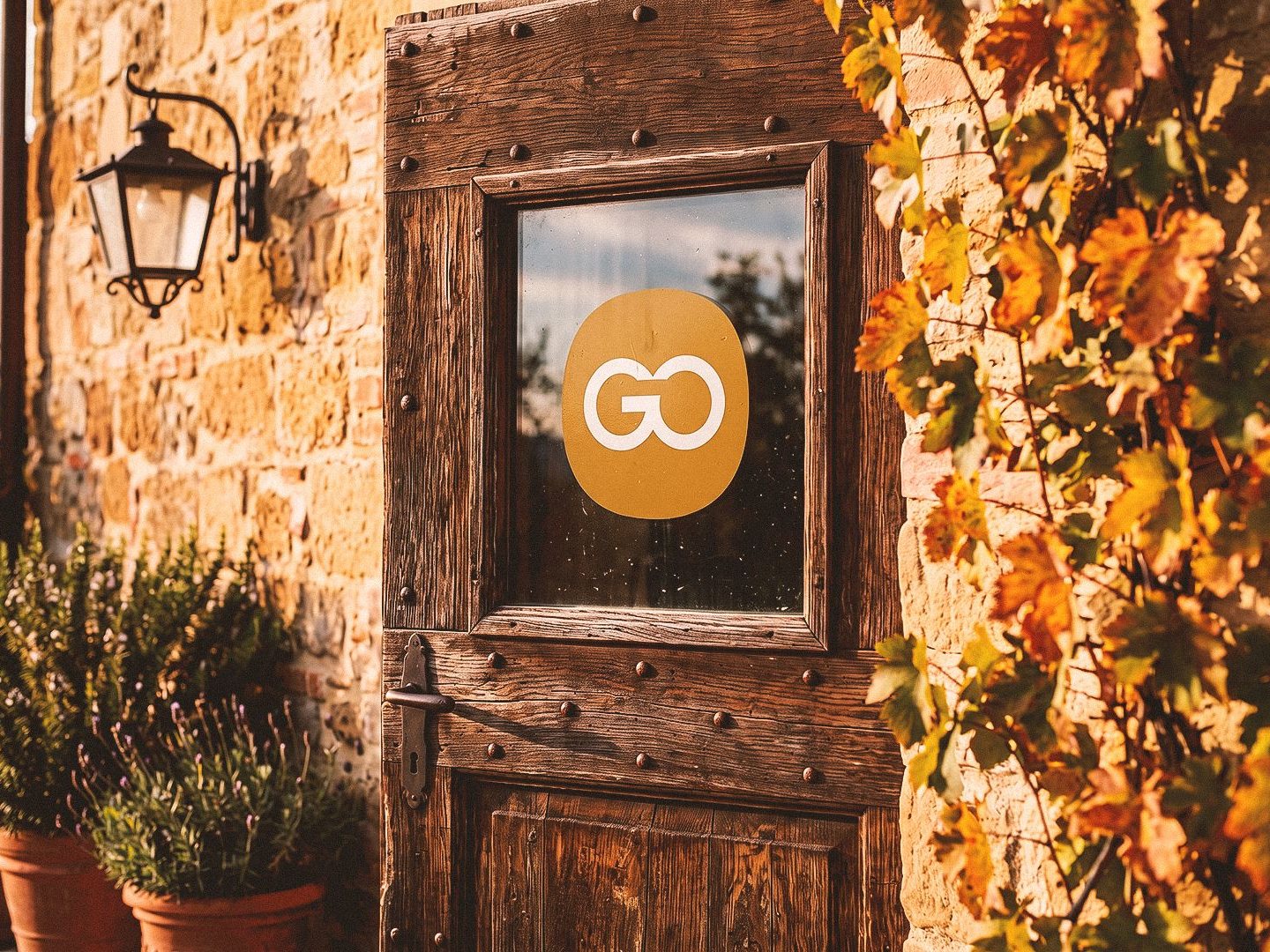 The old wooden door of a stone farmhouse in late sun, a round ochre GO seal on its glass pane, a lantern and a vine with autumn leaves beside it.