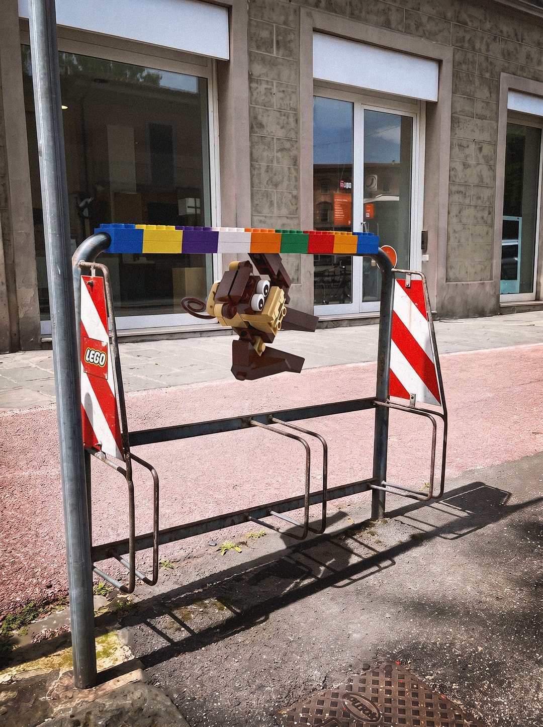 A street bike rack with its top bar covered in a rainbow row of LEGO bricks and a brick-built monkey hanging from it by one arm.