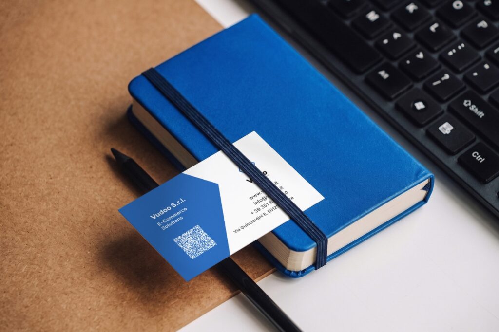 A Vudoo business card tucked under the band of a blue notebook: blue back with the company name and a QR code, white front with the logo and contacts.