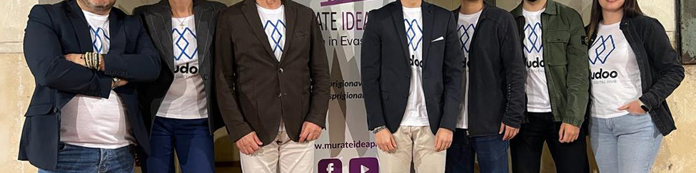 Seven members of the Vudoo team side by side in white t-shirts printed with the stacked logo, in front of a Murate Idea Park banner.